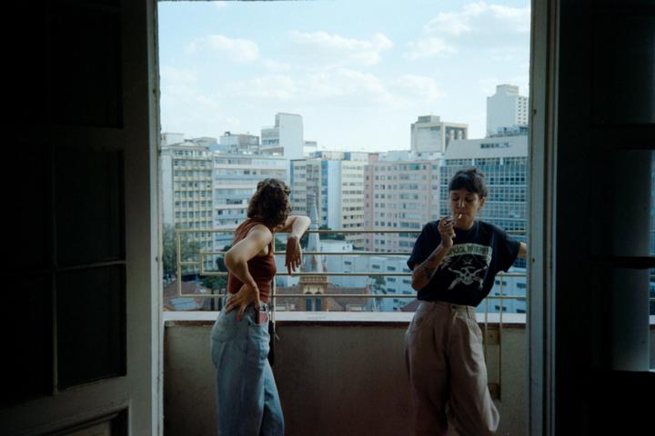 Two women are standing on a balcony overlooking a city with tall buildings under a bright sky.