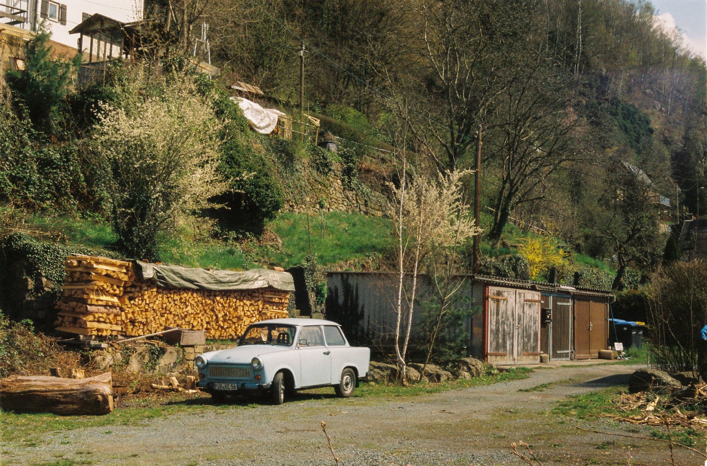 A light blue car is parked on a gravel road next to a large stack of firewood and several sheds, with a steep, tree-covered hillside in the background.