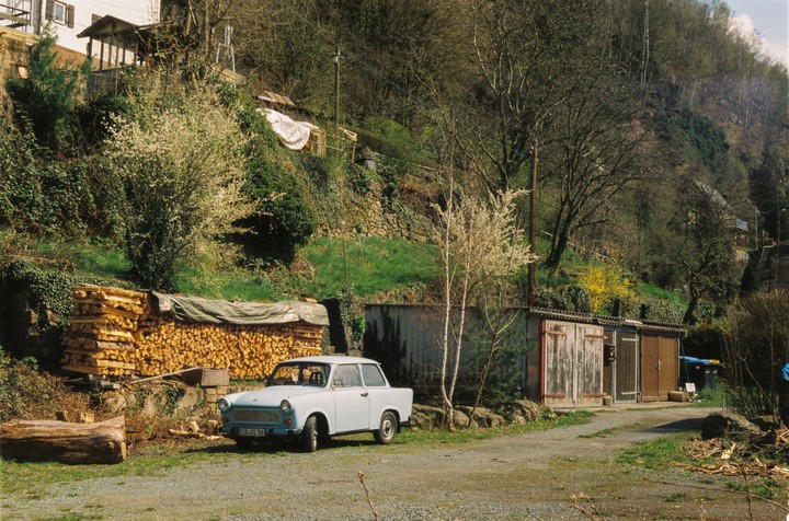A light blue car is parked on a gravel road next to a large stack of firewood and several sheds, with a steep, tree-covered hillside in the background.