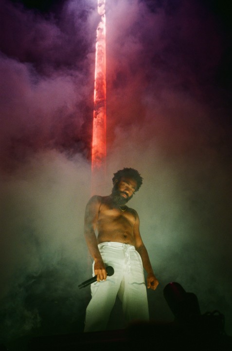 A man with a beard and no shirt holds a microphone on a stage with a red light beam and smoke.