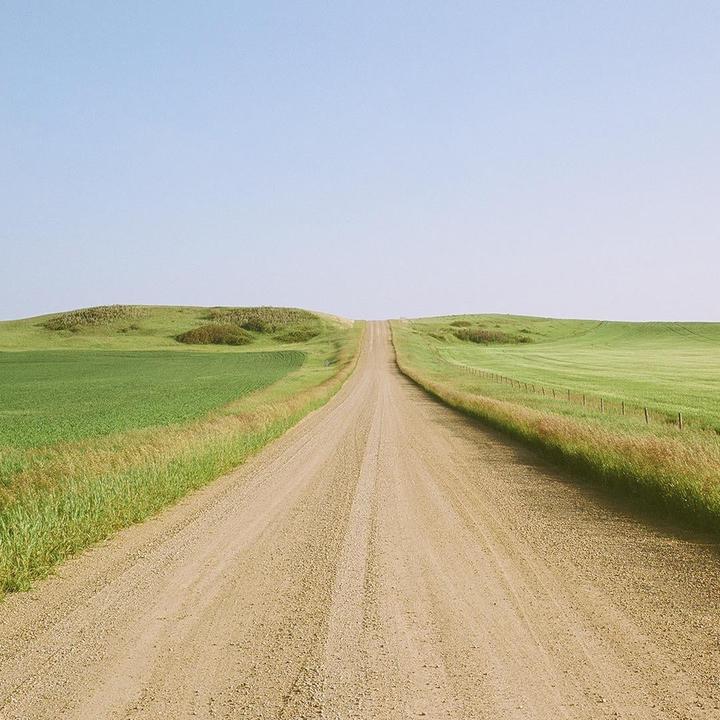 A long gravel road stretches into the distance, flanked by green fields and rolling hills under a clear sky.