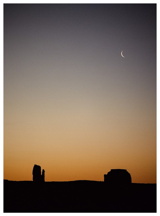 A crescent moon hangs in a gradient sky above silhouetted rock formations at sunrise.