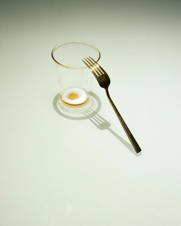 A golden fork rests on the rim of a glass containing a small amount of liquid with a yellow center.