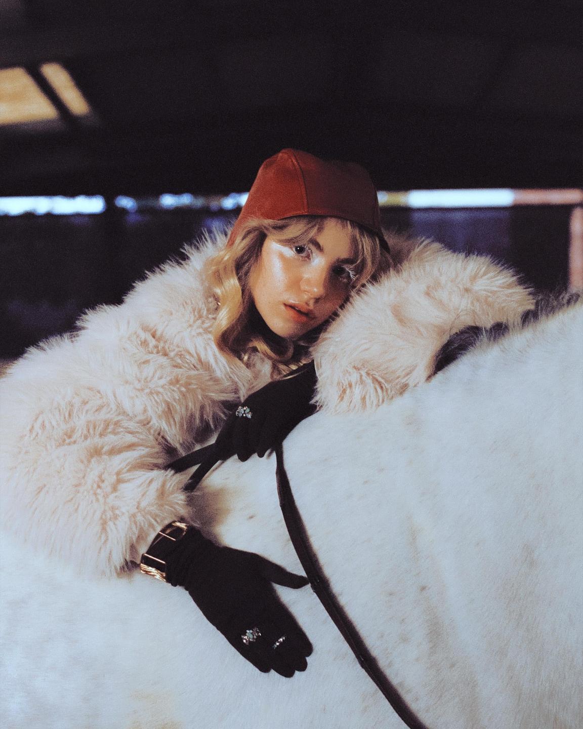 A woman in a fur coat and red hat is leaning on a white horse.