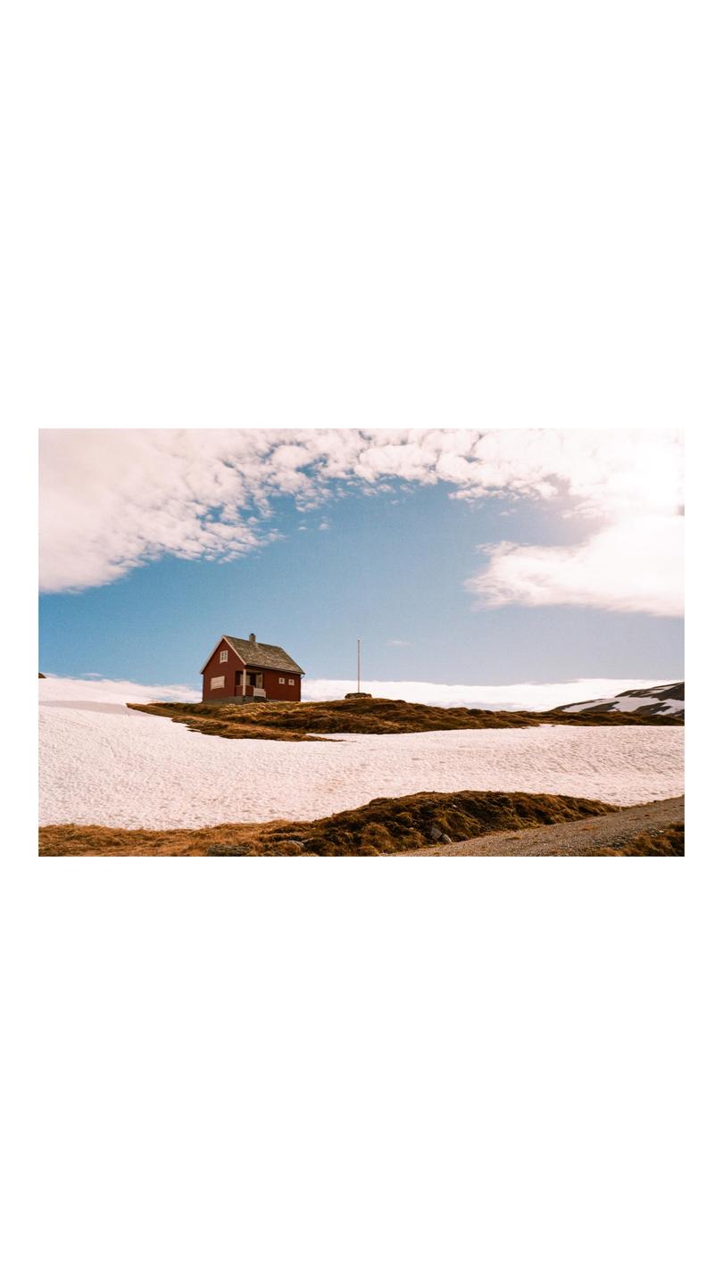 A red house stands on a hill with patches of snow and dry grass under a partly cloudy sky.
