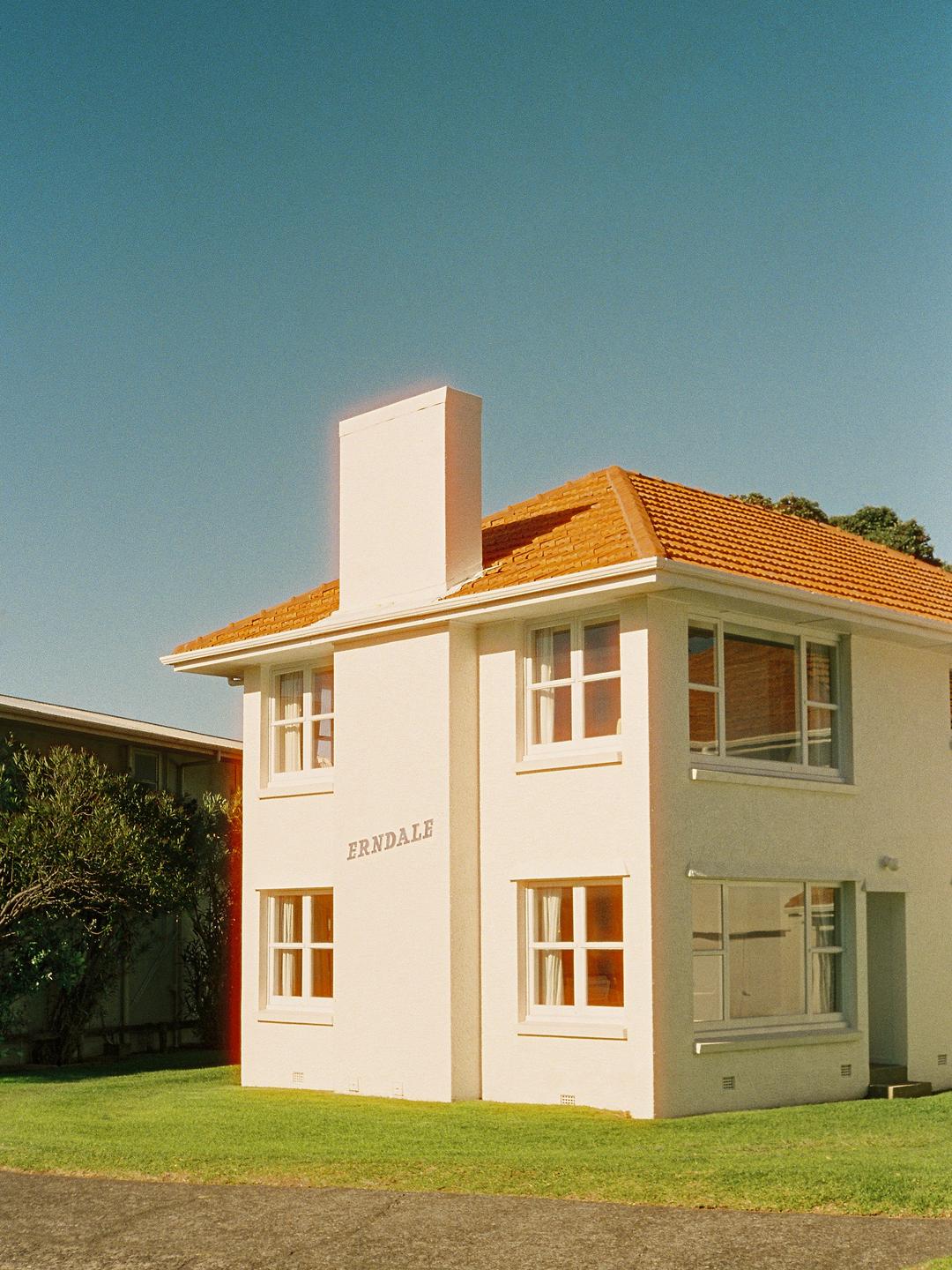 A cream-colored house with a red tile roof and a prominent chimney stands on a green lawn under a clear blue sky.