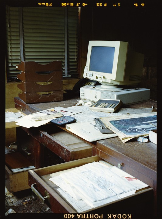 An old computer monitor and tower sit on a dusty desk with papers and office supplies in an abandoned office.