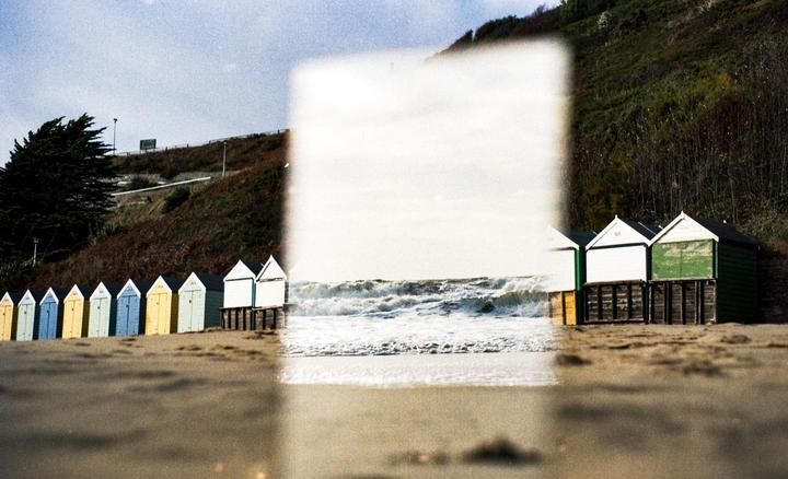 A row of colorful beach huts stands on a sandy beach next to a hill, with waves crashing in the background.