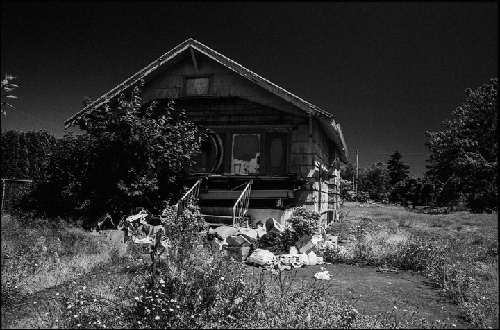 An abandoned house with a porch full of trash is surrounded by overgrown grass and trees.