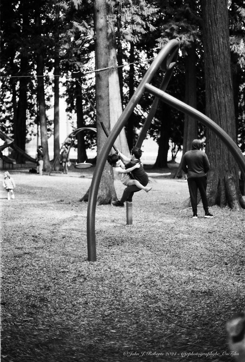 Two children play on a swing set in a park while an adult stands nearby.