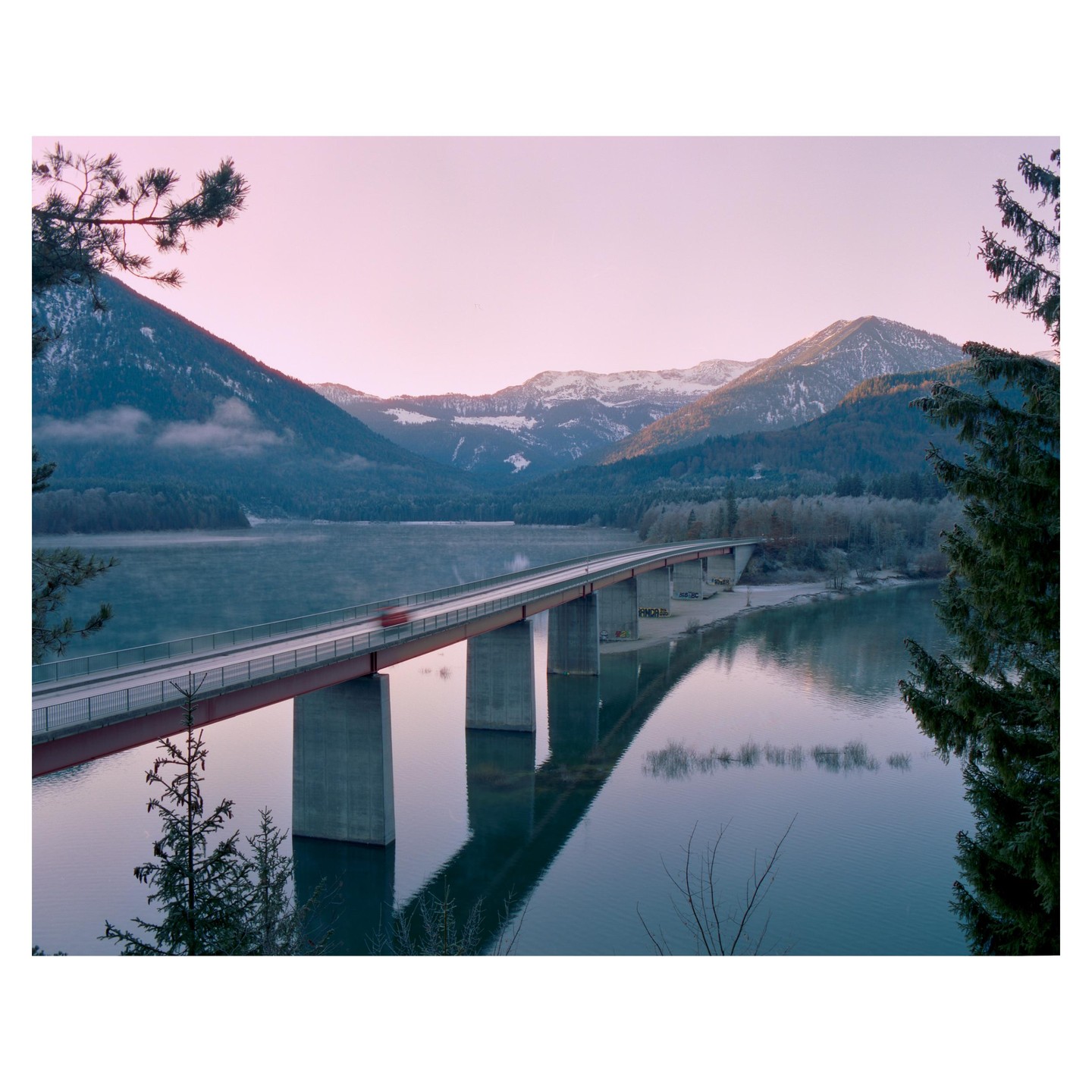 A bridge spans a lake with mountains in the background under a pink sky.