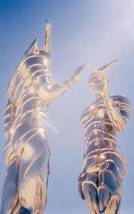 Two shiny, metallic, humanoid sculptures with pointed features stand against a clear blue sky.
