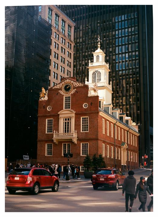 A red brick building with a white cupola and clock tower stands among modern skyscrapers, with cars and people on the street below.