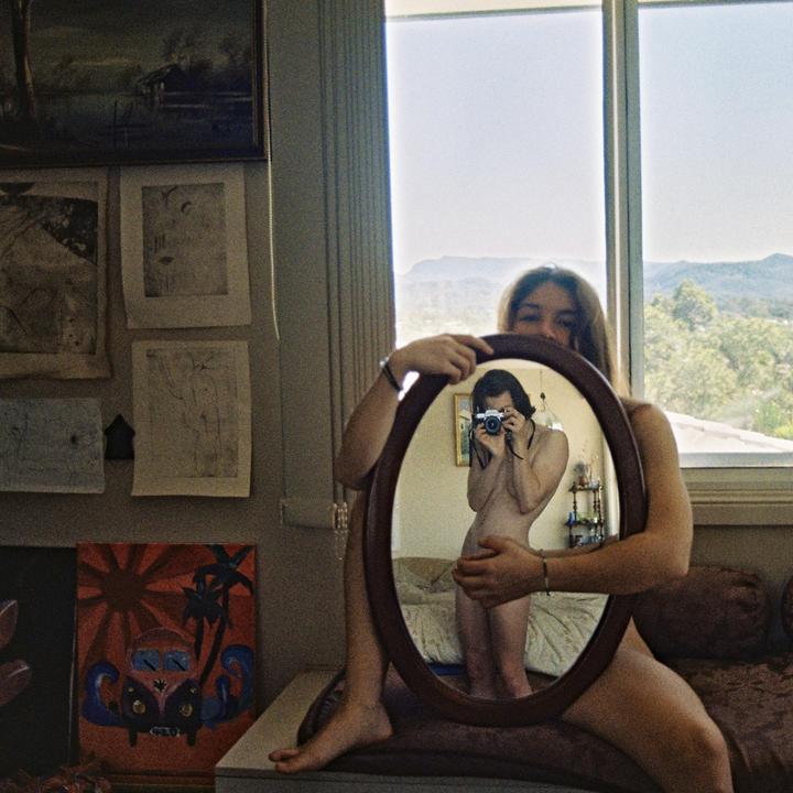 A woman holds an oval mirror reflecting her nude body and a camera, with a window and landscape visible in the background.