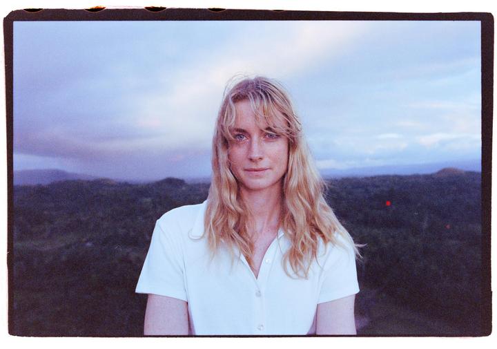 A woman with long blonde hair and a white shirt stands in front of a landscape of hills and a cloudy sky.