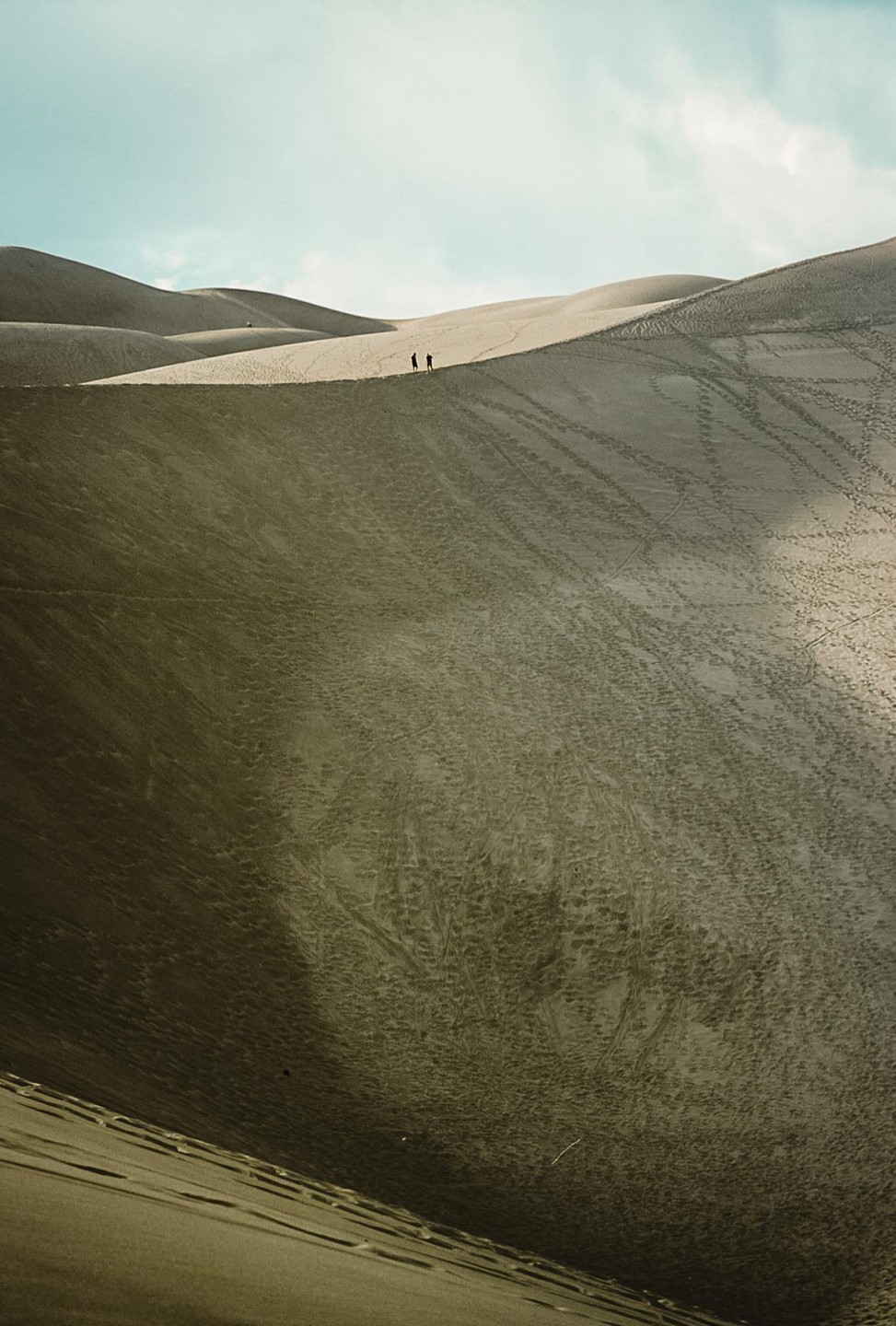 Two people walk along the ridge of a sand dune under a bright sky.