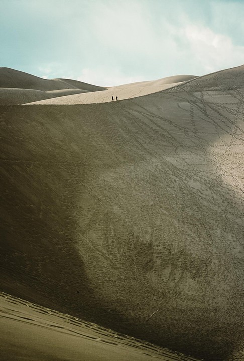 Two people walk along the ridge of a sand dune under a bright sky.