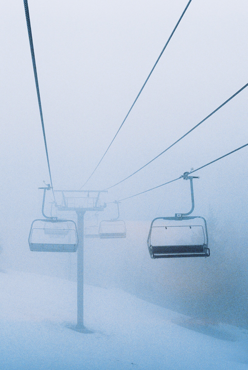 Empty ski lift chairs hang over a snow-covered slope on a foggy day.