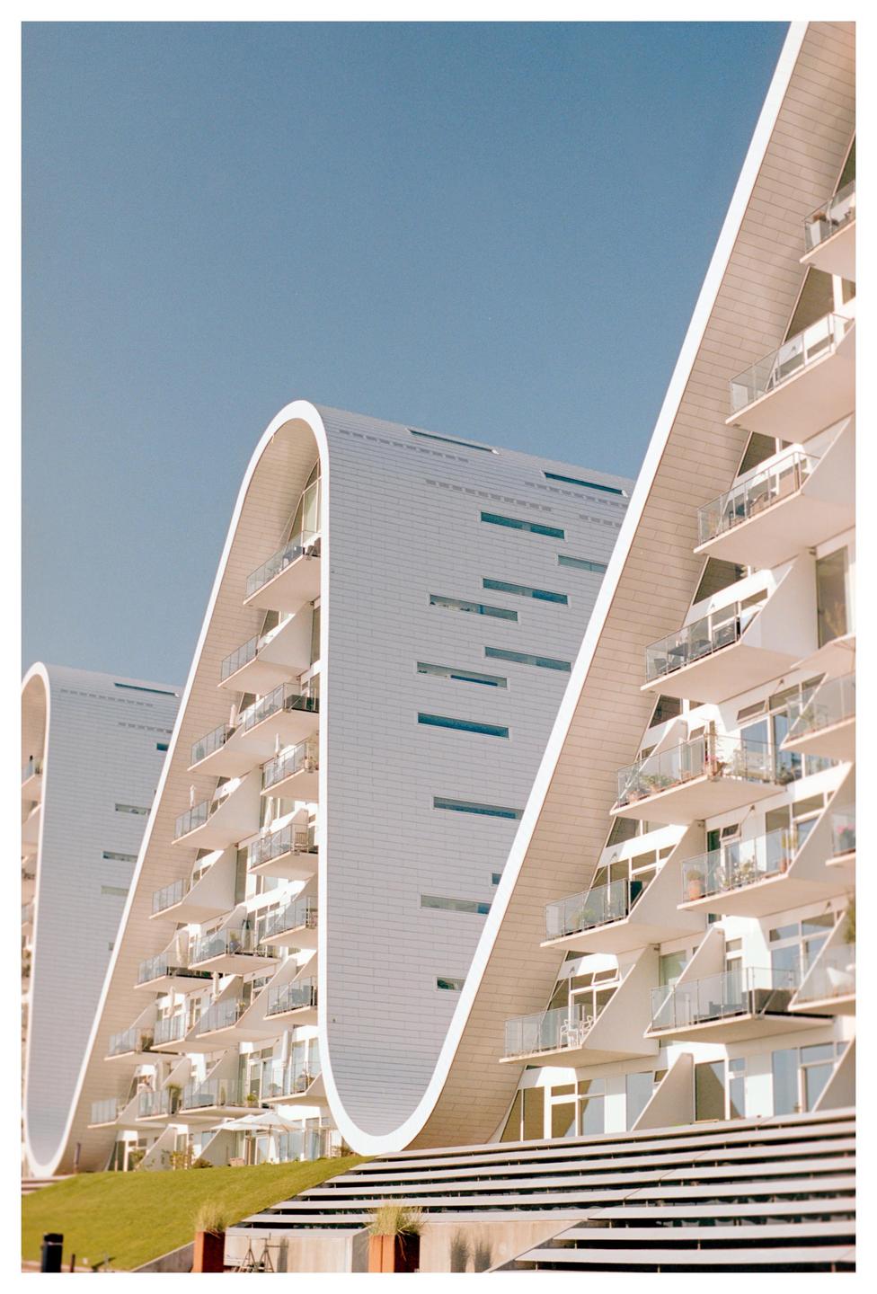 Three modern white buildings with curved roofs and balconies are visible under a clear blue sky.