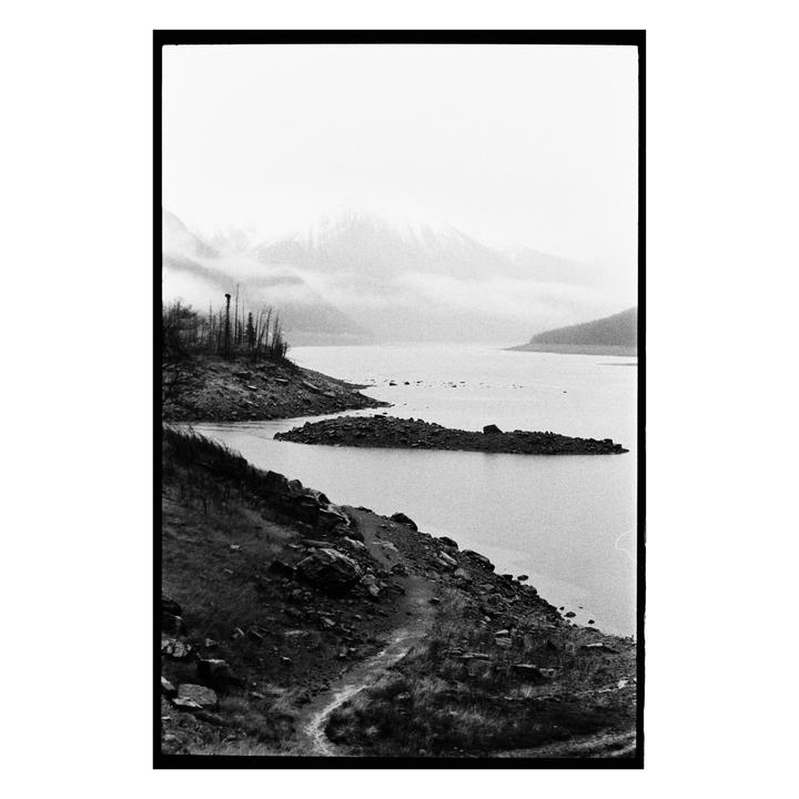 A winding path leads down to a lake with rocky shores and snow-capped mountains in the distance.