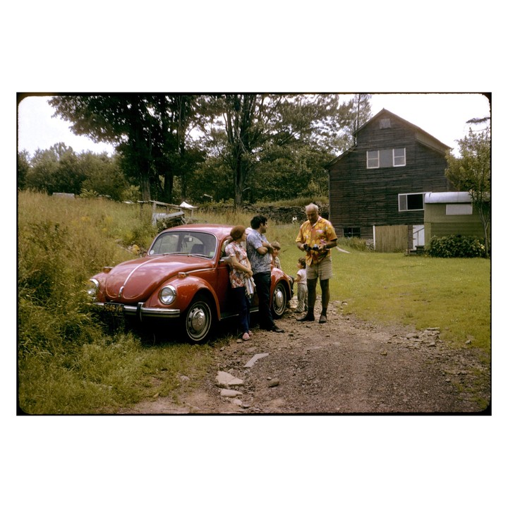 A family stands next to a red volkswagen beetle on a dirt road in front of a wooden house.