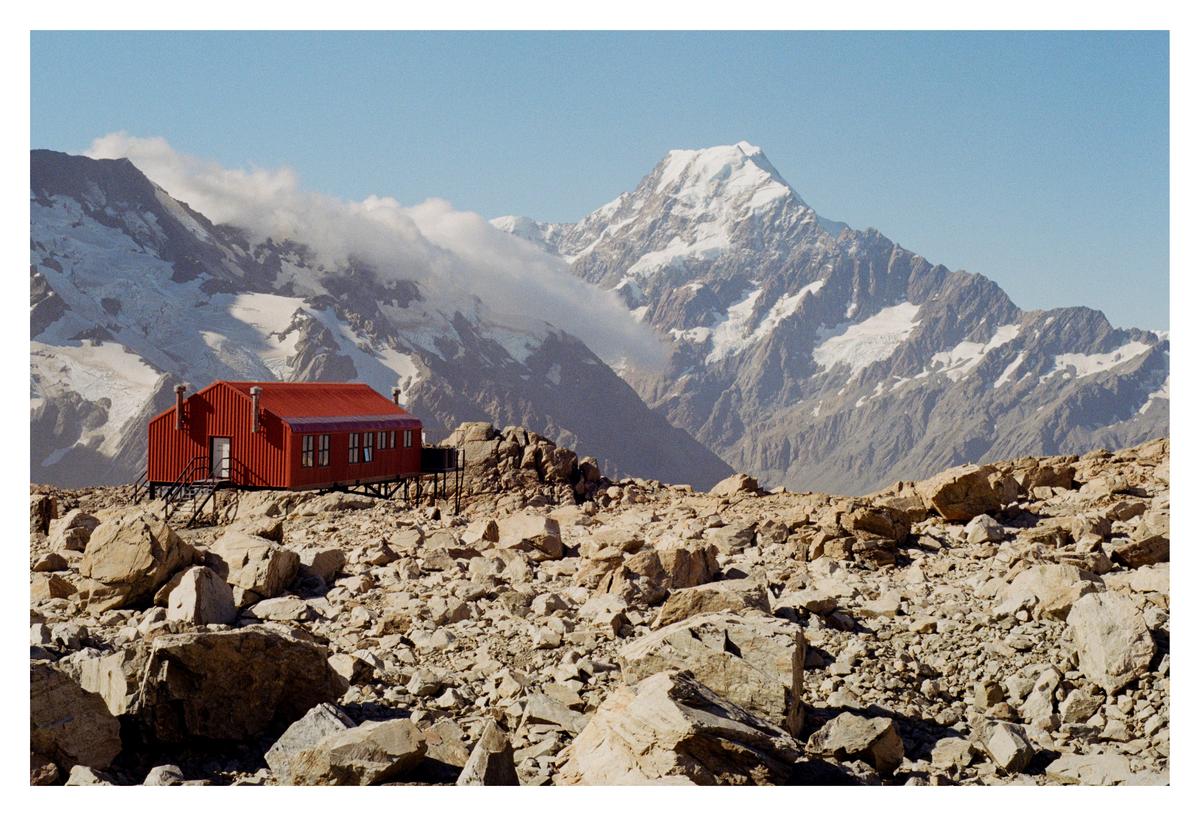 A red hut stands on a rocky, barren landscape with snow-capped mountains in the background under a clear sky.