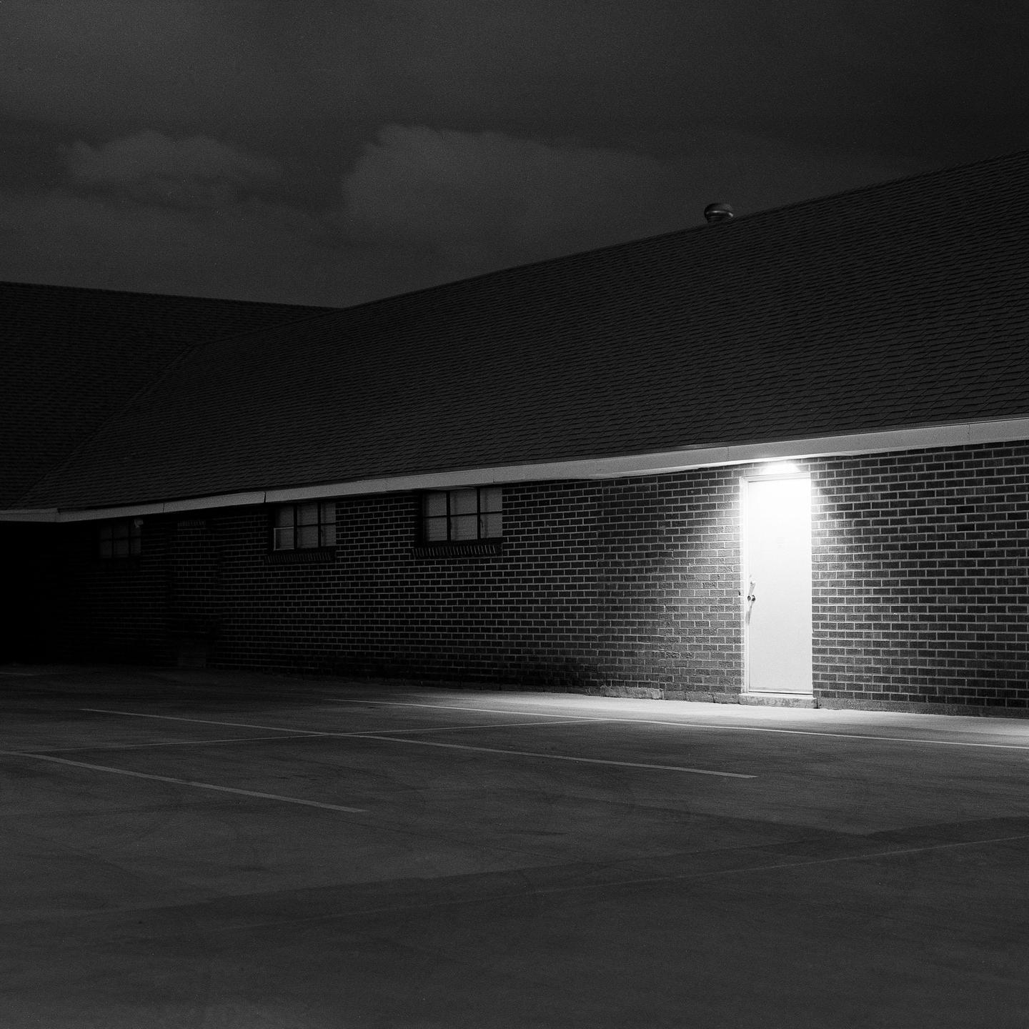 A brightly lit door stands out against a dark brick building with a parking lot in the foreground.