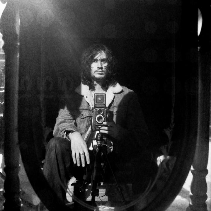 A man with long hair and a denim jacket holds a camera on a tripod in a mirror self-portrait.