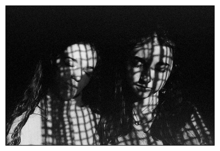 Two women are illuminated by window light creating a grid shadow pattern on their faces and bodies.
