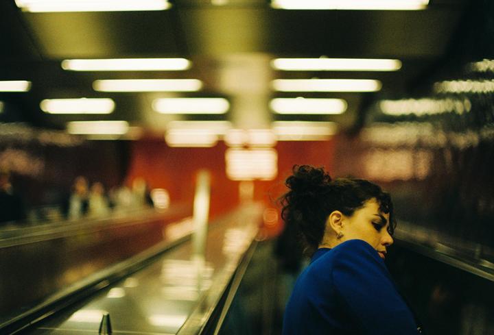 A woman with dark curly hair in a blue top stands on an escalator with blurred lights in the background.