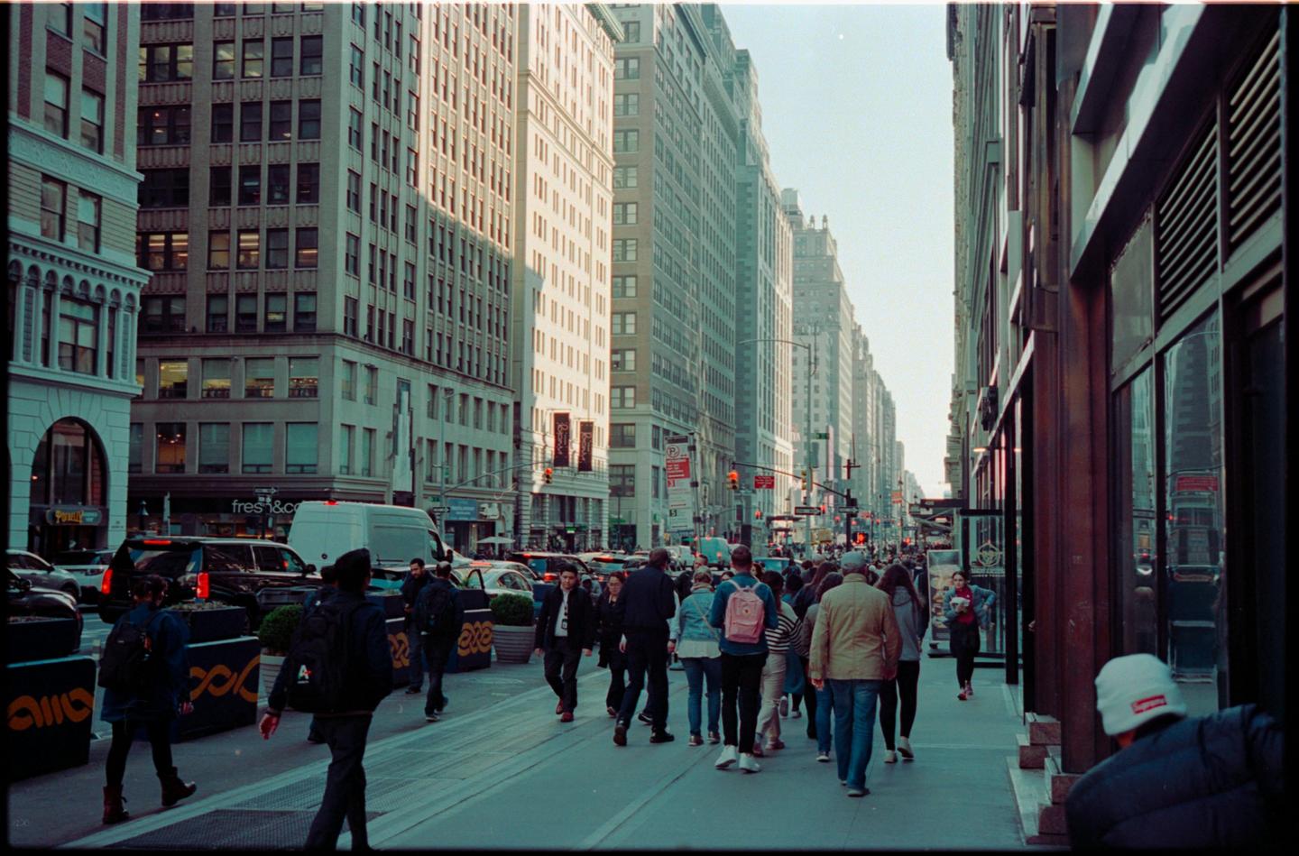 A busy street scene shows pedestrians walking on a sidewalk next to tall buildings and parked cars.