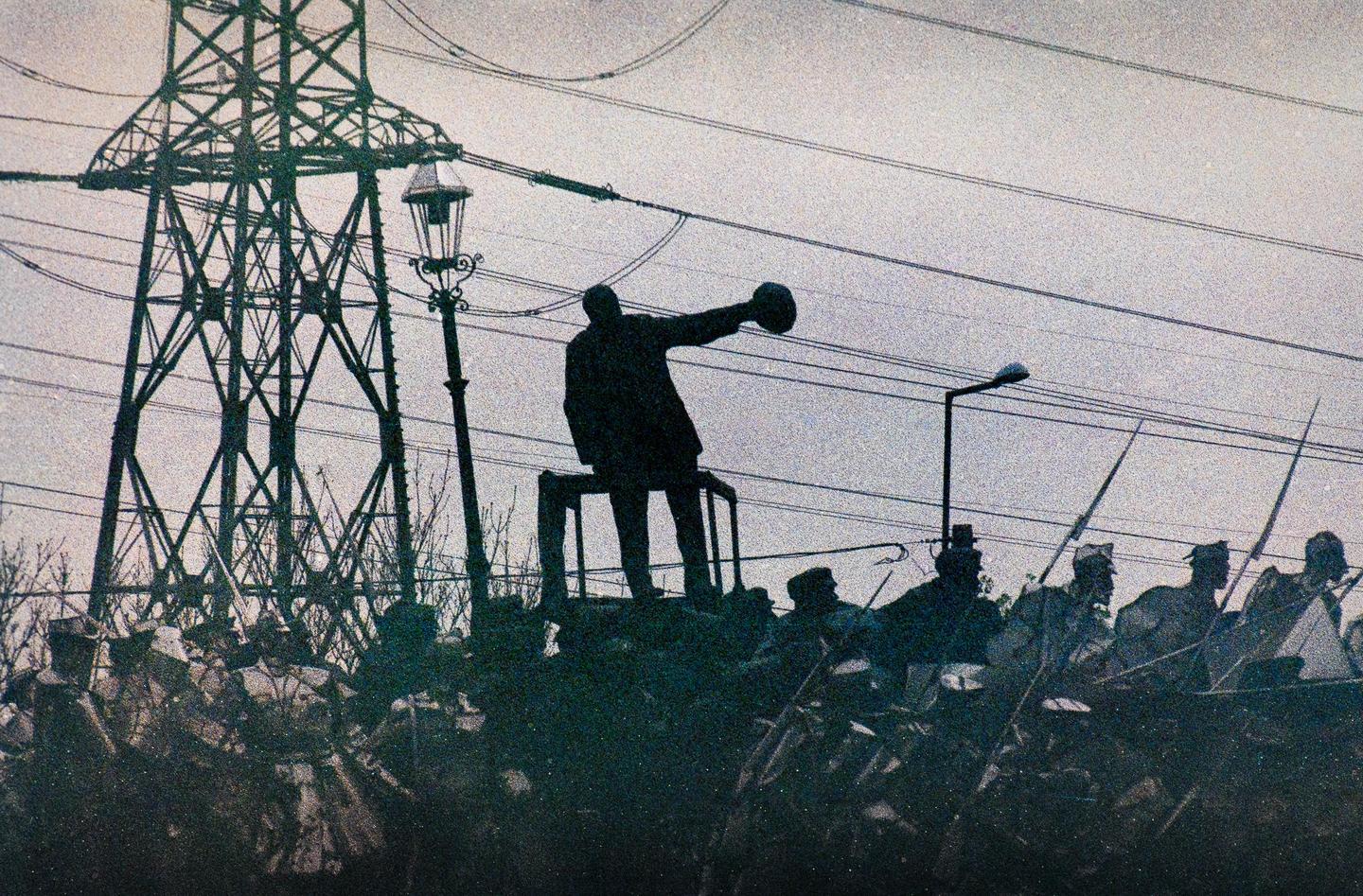 A statue of a man stands on a pedestal with a crowd of people below, next to a large metal tower and power lines.