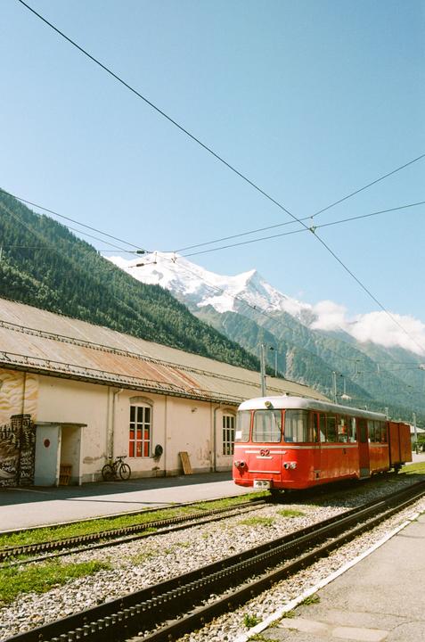 A red train is parked on tracks next to a building with a snow-capped mountain and forest in the background.