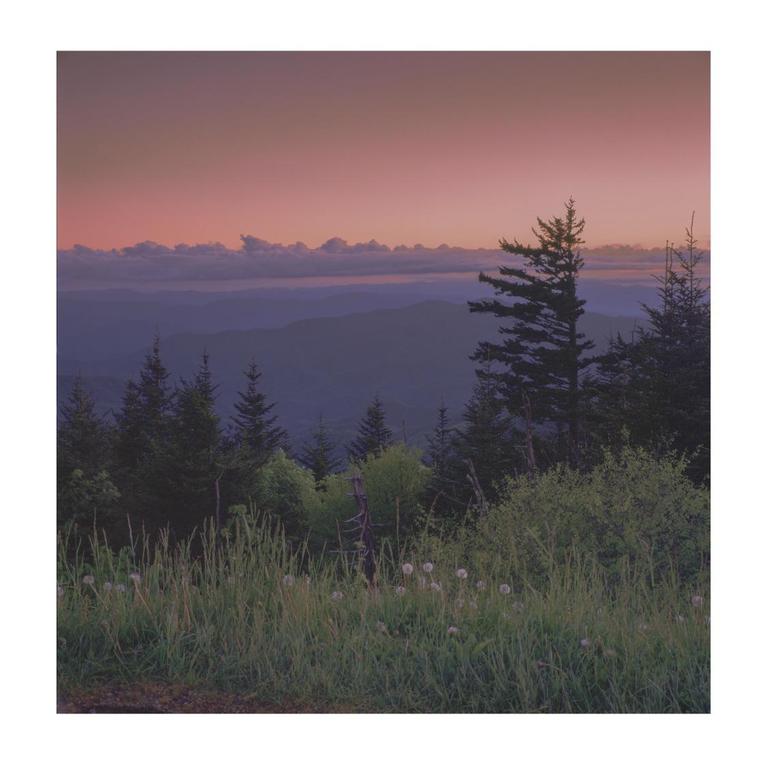 A mountain landscape at dusk shows a pink sky over layers of blue mountains and green trees.