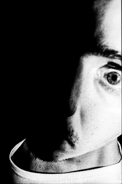 A man's face is partially visible in a high-contrast black and white close-up, with one eye wide open and the other side of his face obscured by shadow.