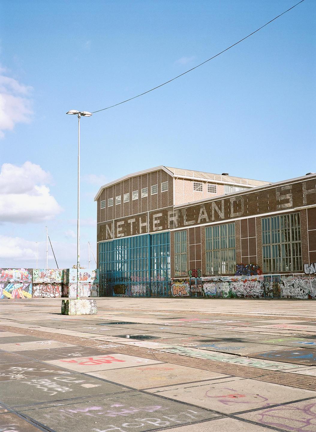 A large brick building with the word "NETHERLANDS" written on its side stands next to a concrete area covered in graffiti under a blue sky.