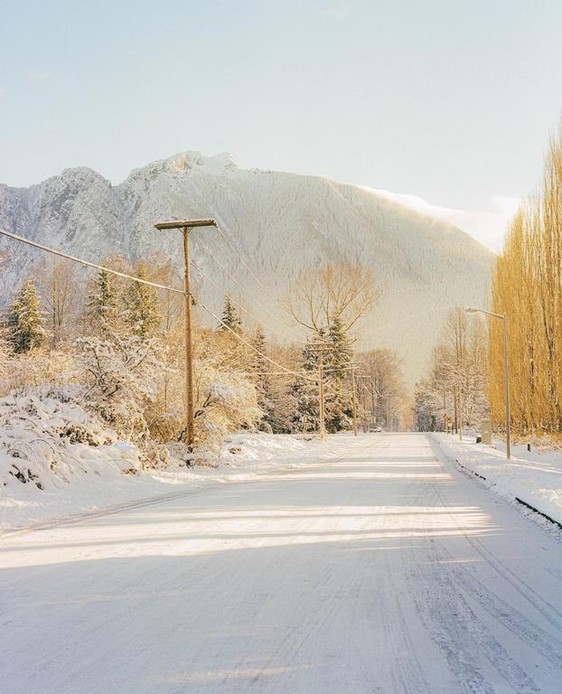 A snow-covered road leads toward a mountain under a bright sky, with trees and utility poles lining the sides.