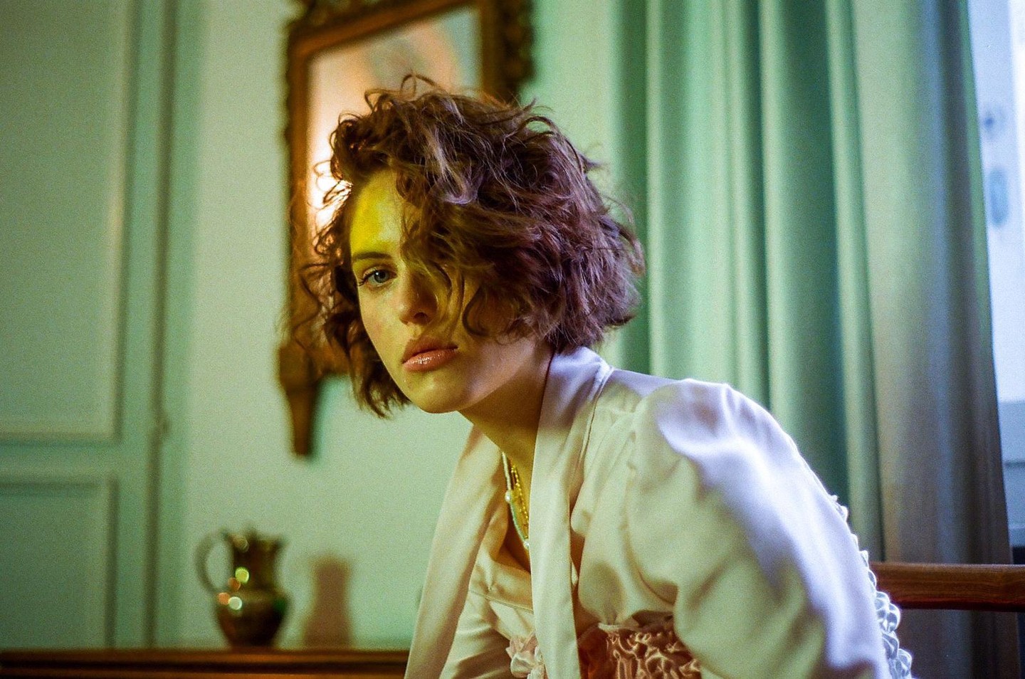 A woman with short, curly hair wears a pink satin robe and looks at the viewer.