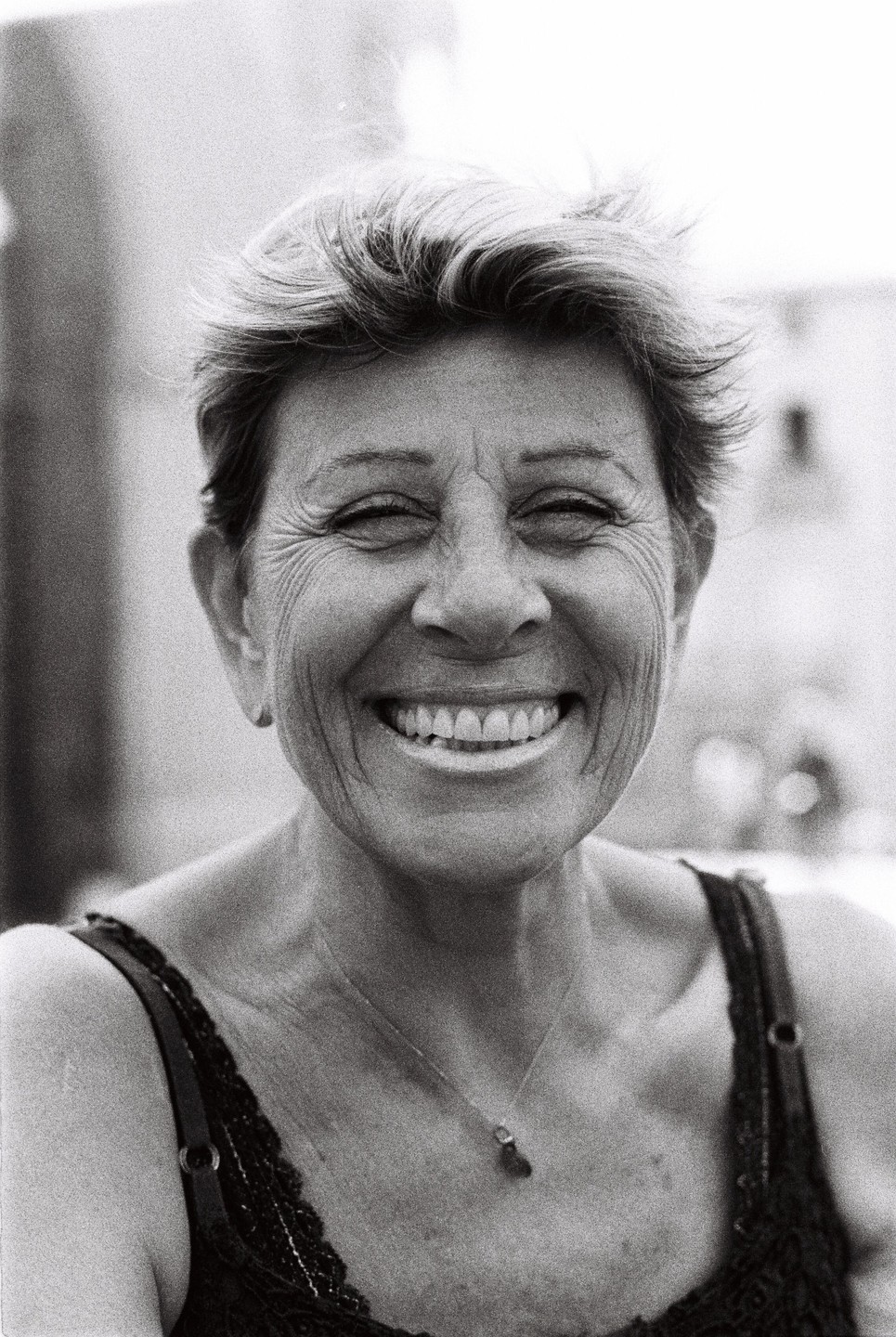 A woman with short hair smiles broadly at the camera, wearing a dark top with a delicate necklace.