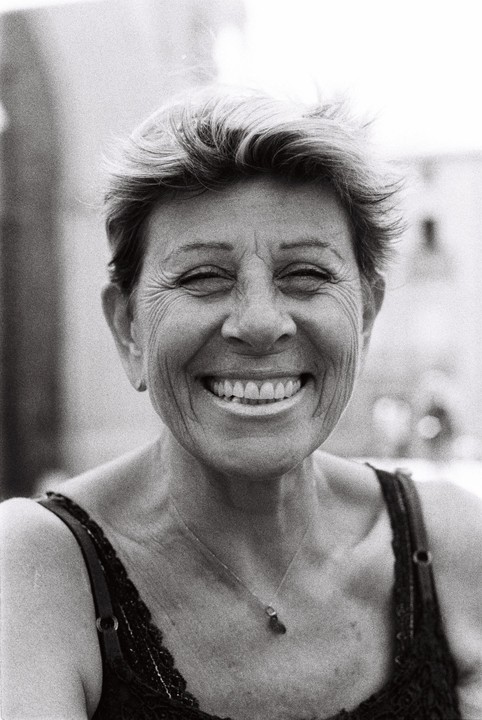 A woman with short hair smiles broadly at the camera, wearing a dark top with a delicate necklace.
