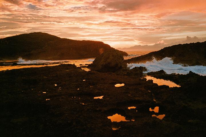 Waves crash against a rocky coastline under a dramatic orange and pink sunset sky.
