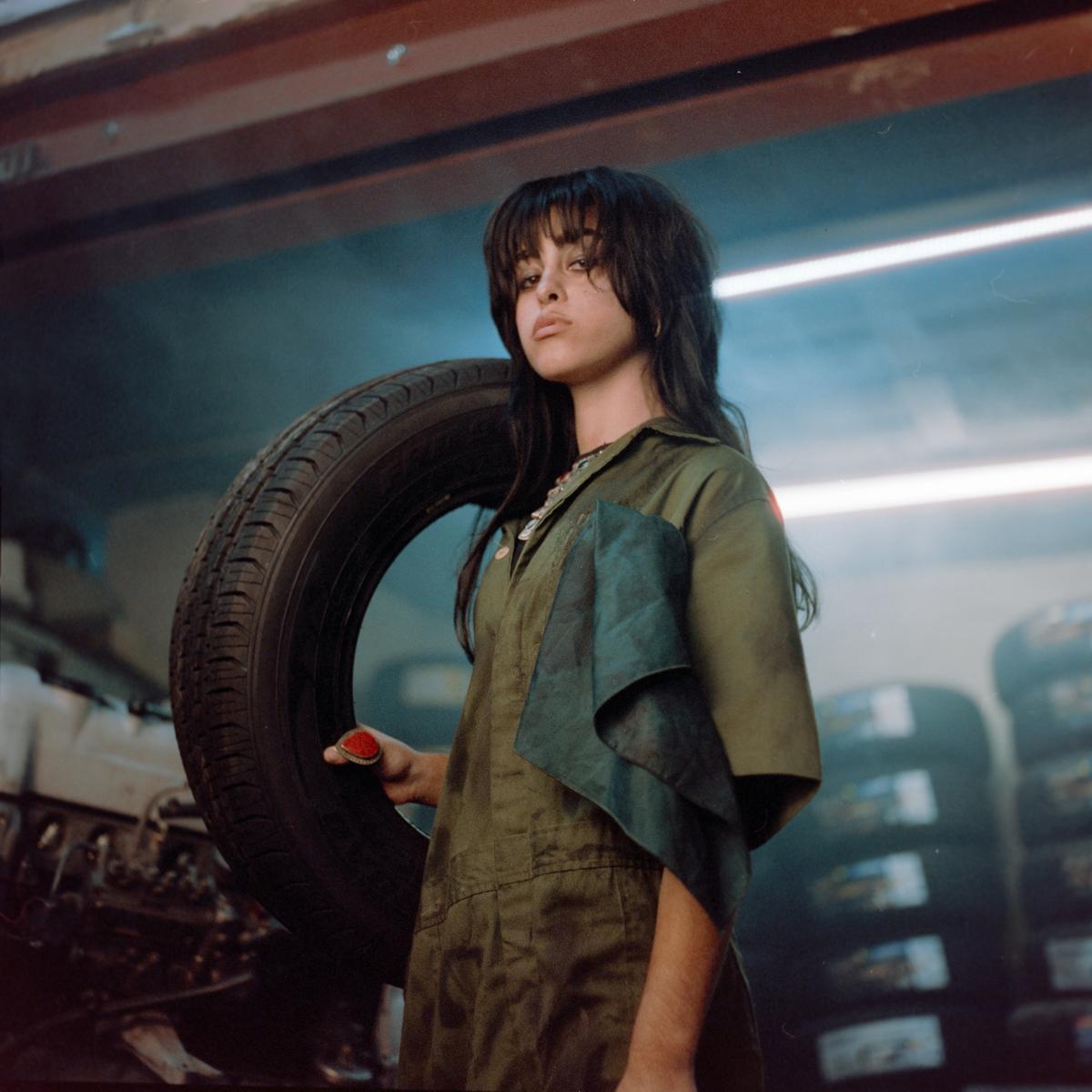A woman in a jumpsuit holds a tire in a garage.