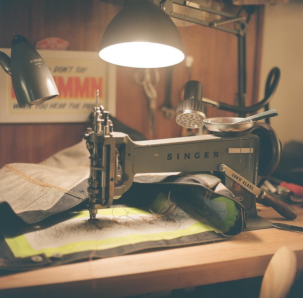 A Singer sewing machine is illuminated by a desk lamp as it stitches a piece of fabric on a wooden table.