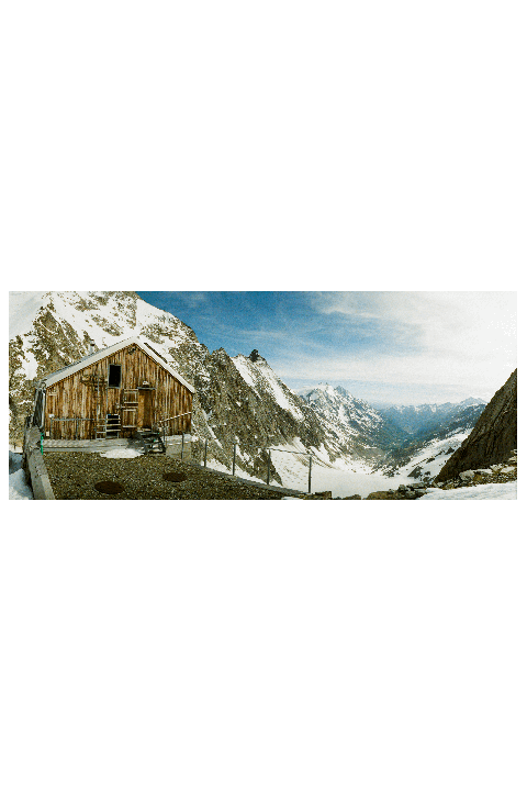 A wooden cabin sits on a rocky ledge overlooking a snow-covered mountain valley.