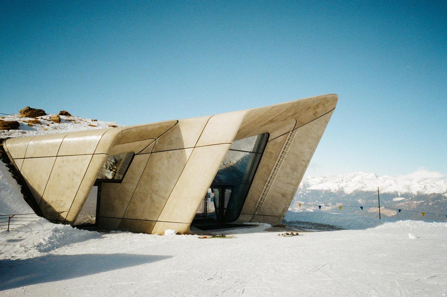 A modern building with angular concrete walls and large windows stands in a snowy landscape with mountains in the background.