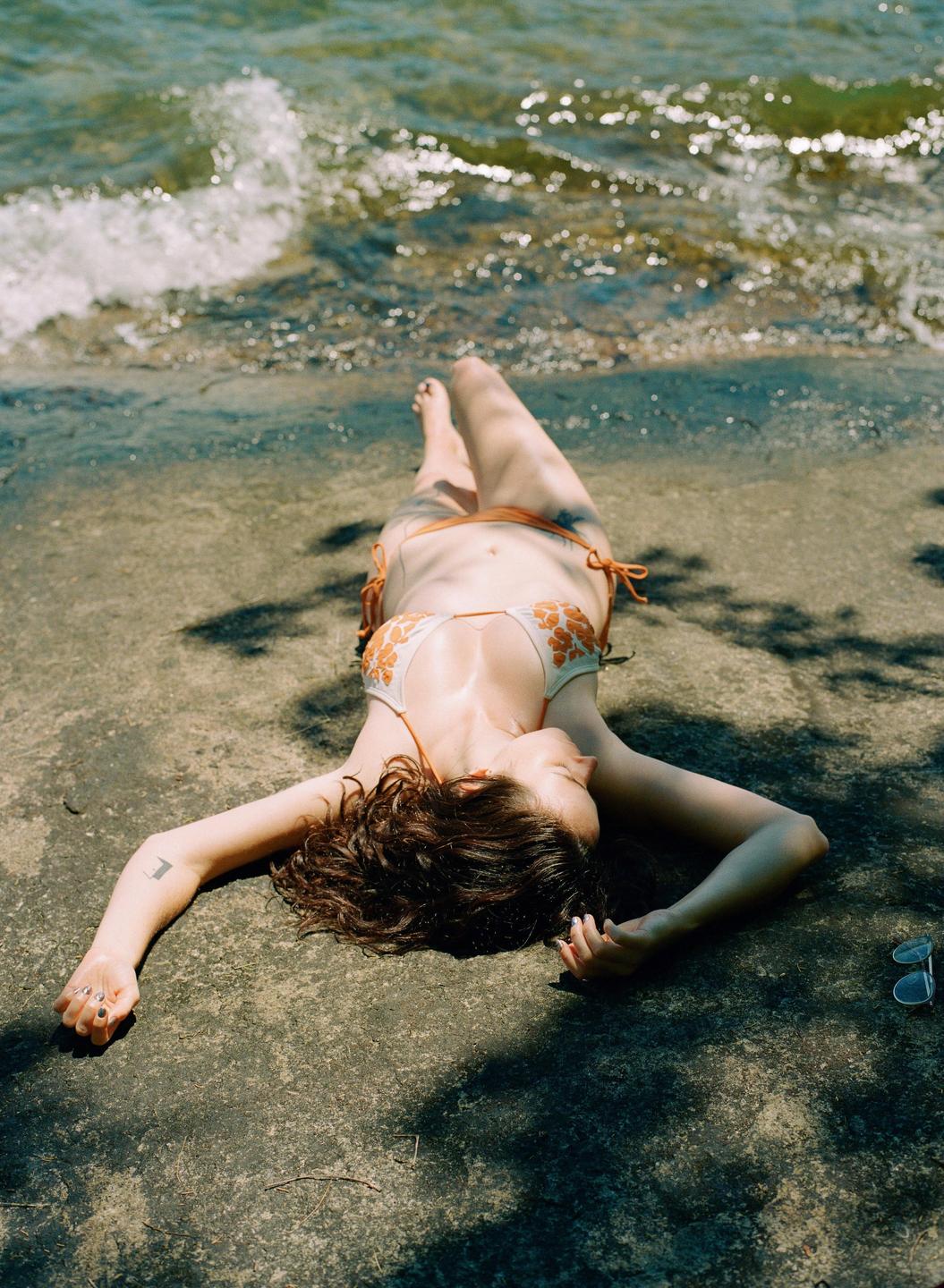 A woman in a bikini lies on a rocky shore next to the water.