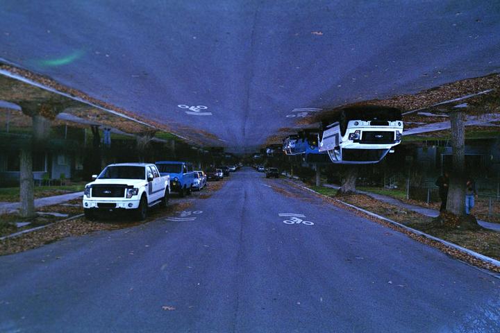A street with parked cars is shown with an inverted, mirrored image of the same street above it, creating a surreal effect.