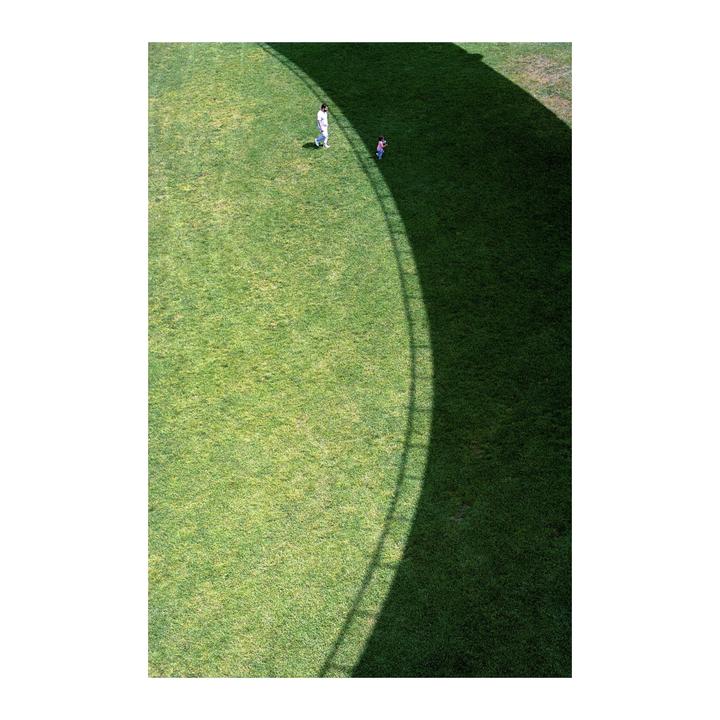 A man and a child walk on a grassy field with a curved shadow.