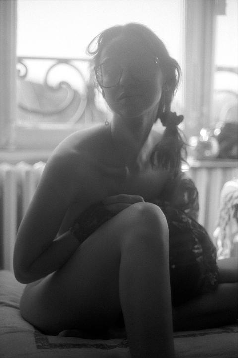 A woman with glasses sits on a bed, backlit by a window.