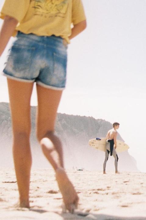 A person in denim shorts walks on a sandy beach towards a surfer holding a surfboard.